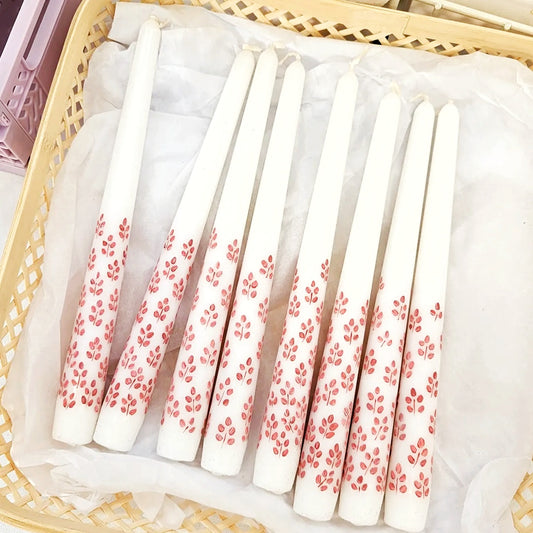 There are 8 hand-painted tapered dinner candles laid out in a wicker style basket with white tissue paper underneath them. The candles are white and they have hand painted clusters of leaves in a park pink. The botanical clusters go about half way up the candles.