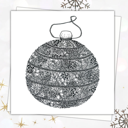 Image shows a Christmas card with an envelope tucked in behind it. On the front of the card there is an illustration of a black and white Christmas bauble on a little string. The pen illustration is made up a mixture of leaves, holly, and dots. There are some Christmassy elements laid out around the background of the image around the festive card.