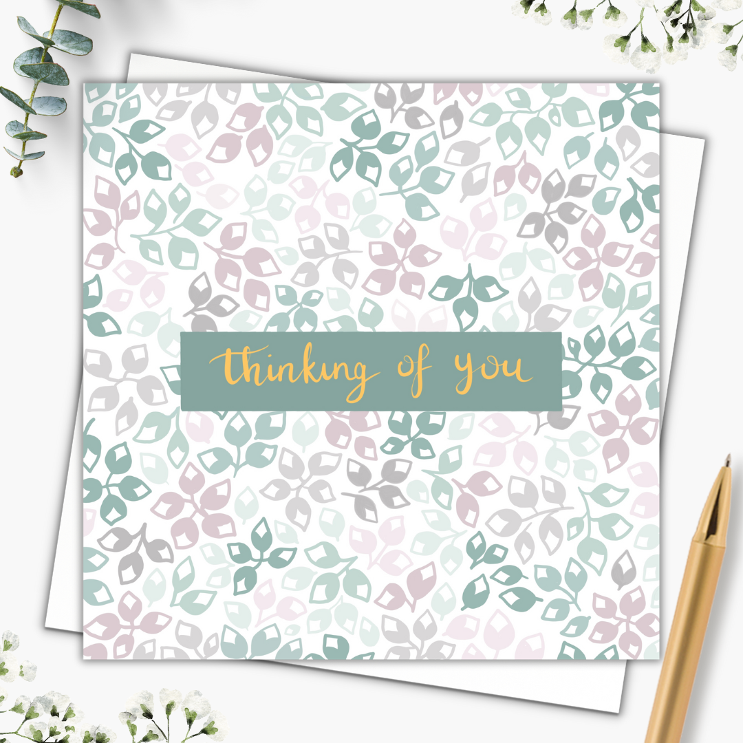 Botanical greeting card with leafy buds of different colours. The colour palette is various shades of green, grey and dusty pink. In the centre of the card are the words "thinking of you" in gold writing on a dark green background. The card is set with an envelope behind it and a gold pen beside it.