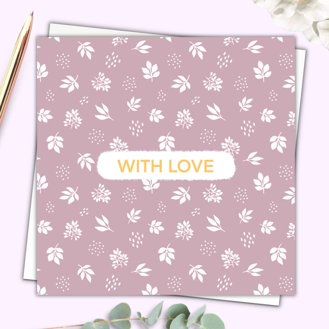 Botanical themed greeting card with the words "With Love" in the centre. The words are written in a simple gold font with a small white surround. The rest of the card is a botanical pattern of white leaves and petals set on a dusty dusk pink. The card is set on top of a plain white envelope beside a gold pen.