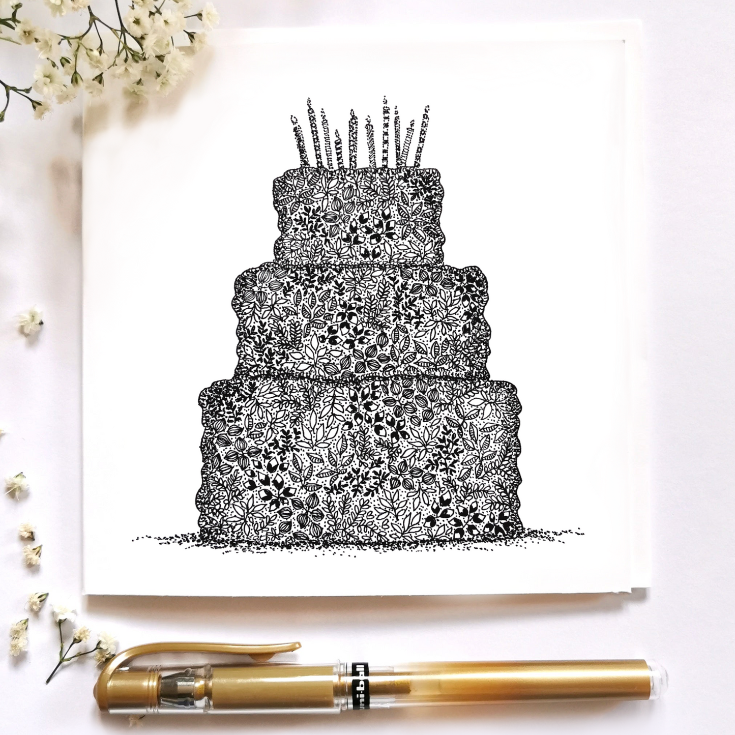 Image shows three tier birthday cake illustration drawn mostly up of a variety of flowers. 10 Candles are places on top of the cake with little dots at bottom of the cake to showcase the height of the cake. Image is places on top of a cream background with white flowers top left and bottom left corner and a gold pen at the bottom of the page.