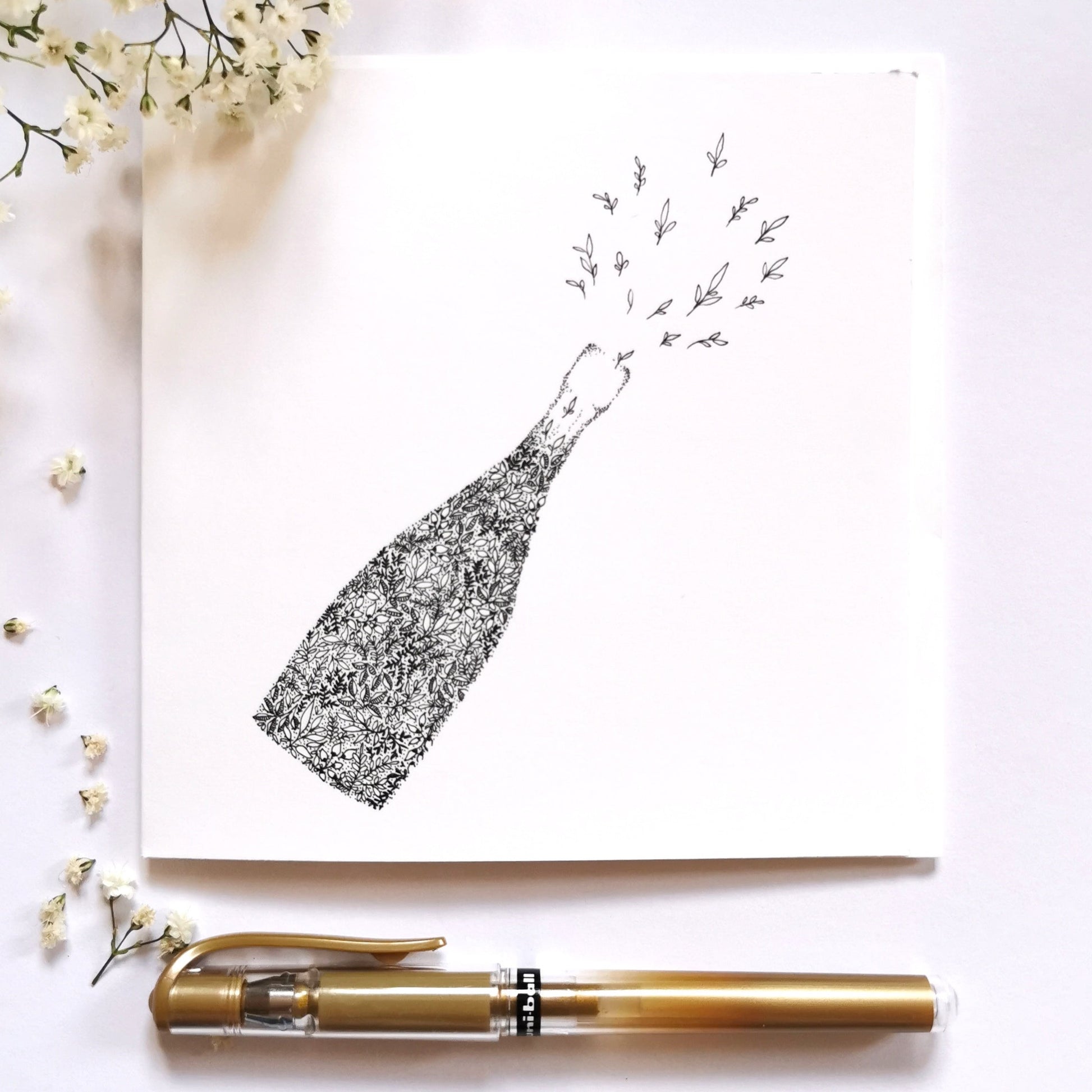 Image shows illustration of champagne bottle made entirely of black and white floral drawings. Bottle is slanted towards the right as if it had just been popped open with tiny flowers spritzing out the bottle as if it was the foam of a champagne bottle popping. Image is set on a cream table with a gold pen at the bottom of the image and white flowers on the left hand side of the image to show what illustration would look dressed up
