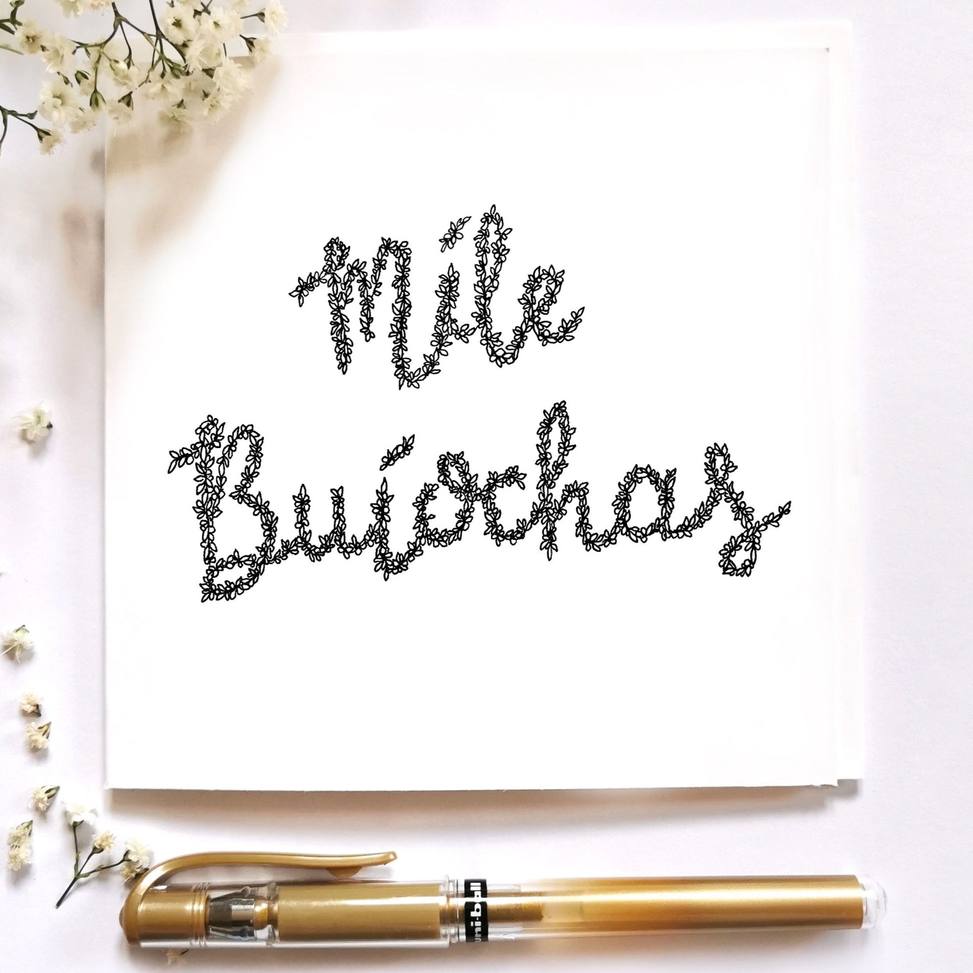 Image shows illustration of the Irish saying Míle Buíochas meaning Thanks A Million. illustration is drawn from petals flowers and a variety of plants. Image is entirely black. Illustration is set on a white card that's laid on a cream background with gold pen and white floral arrangements to dress up.
