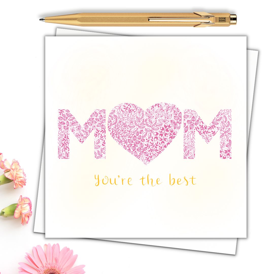 Image shows MUM YOURE THE BEST card. MUM has a pink heart in place of the U and YOURE THE BEST is drawn in yellow writing. MUM is drawn in pink writing and taking the fore front of the card. The heart is made from pink floral drawings. The image is laid on a cream surface with a gold pen and pink flowers surrounding it. 