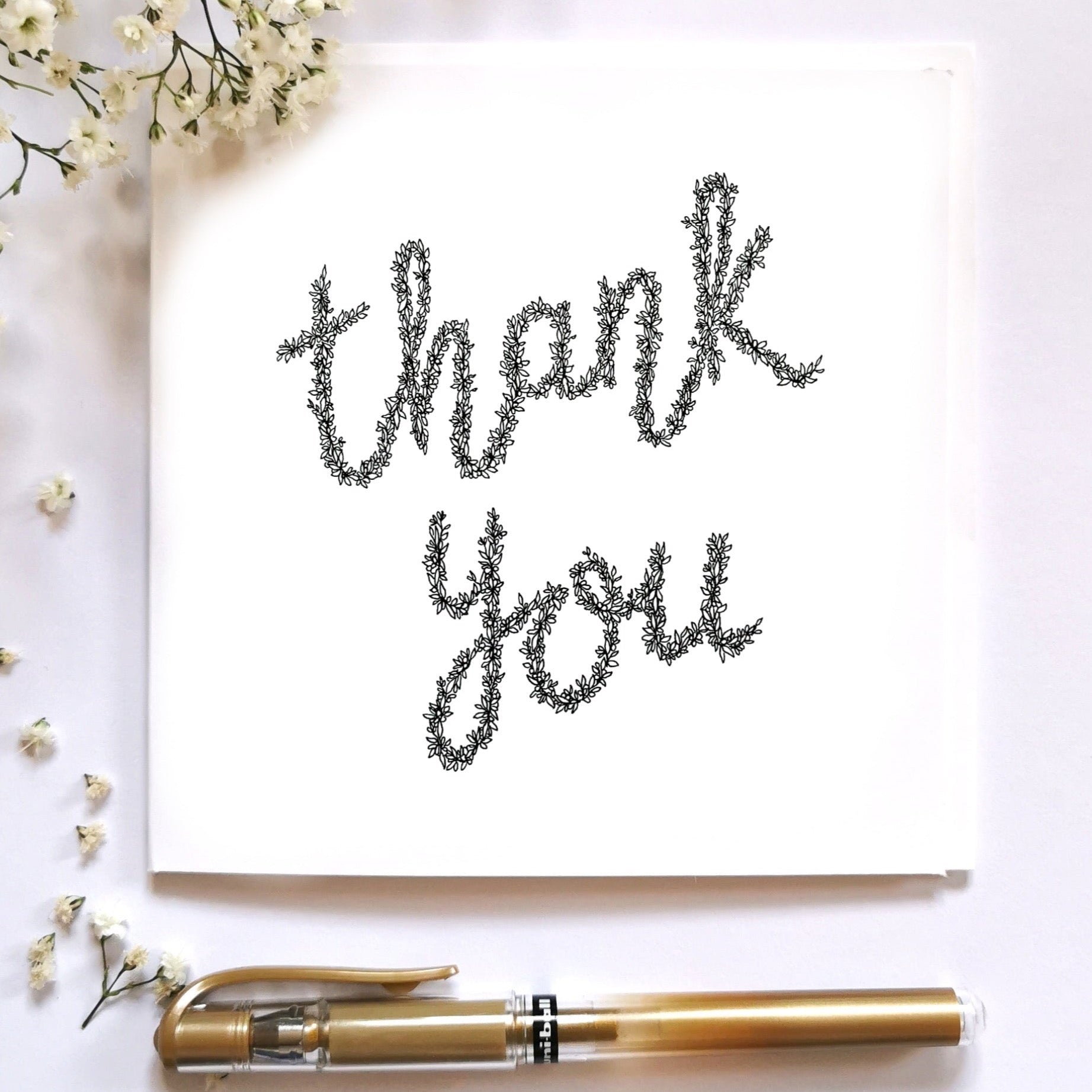 Image shows Thank you card made from black floral drawing. Image laid on cream surface with floral arrangement and gold pen. 