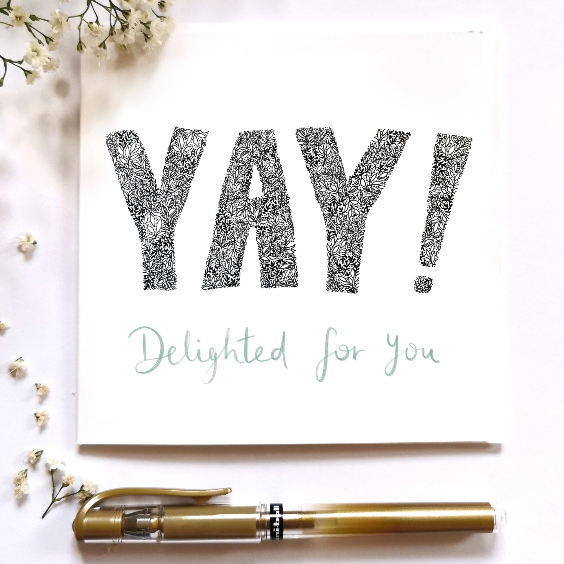 image shows YAY DELIGHTED FOR YOU illustration card. YAY is written in cap block writing drawn from flowers and leaves. and DELIGHTED FOR YOU is written in gold pen. Image is placed on a cream surface with a gold pen and white flowers. 